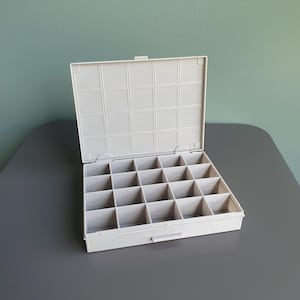 Puede incluir: Una caja organizadora de plástico gris claro con tapa abatible y veinte compartimentos cuadrados individuales. El interior está dividido en un patrón de cuadrícula, proporcionando secciones organizadas para objetos pequeños en el contenedor.