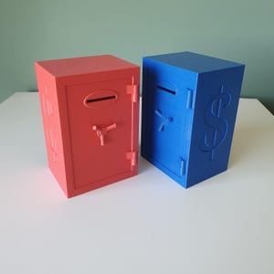 May include: Two colorful piggy banks shaped like safes. One is red, the other is blue. Each has a slot for inserting money, a combination dial, and a door. The blue bank features a large dollar sign on its side.