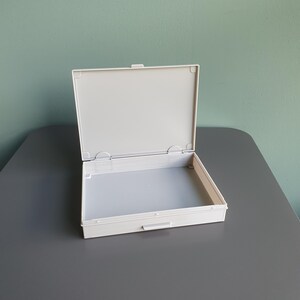 Peut inclure: Une boîte de rangement rectangulaire en plastique blanc cassé avec un couvercle à charnière ouvert. Le récipient présente une base intérieure lisse et plate ainsi qu'un mécanisme de fermeture simple sur le bord avant pour ranger des objets.