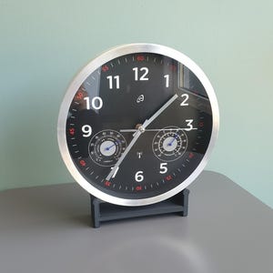 Op de afbeelding: Een ronde wandklok met een zilverkleurige rand en een zwarte wijzerplaat met witte Arabische cijfers. De klok bevat twee kleinere sub-wijzerplaten voor luchtvochtigheid en temperatuur, zilveren wijzers en rode minuutmarkeringen.
