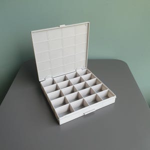 Op de afbeelding: Een vierkante opbergdoos van gebroken wit plastic met een vastzittend scharnierend deksel. De binnenkant is voorzien van een raster van zestien individuele vierkante vakjes in een vier-bij-vier patroon voor het opbergen van items.