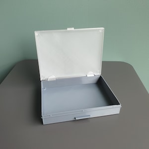May include: A rectangular storage box with a grey base and a hinged white lid. The lid features a subtle diagonal textured pattern across its surface. The box is open, revealing a smooth, flat interior base and a small latch on the front edge.
