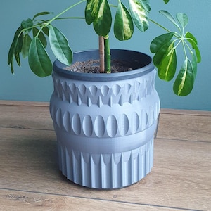 May include: A matte grey cylindrical plant pot featuring a textured exterior with repeating vertical grooves and rounded indentations. The pot has a dark grey inner rim and contains a green plant with variegated leaves extending from the centre.