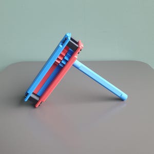 May include: A handheld ratchet noisemaker featuring a bright blue and red plastic frame with a central gear mechanism and a long blue handle. The device is constructed with layered components and black accents, designed to produce sound when rotated.