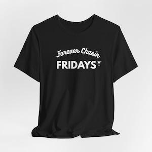 Puede incluir: Camiseta negra con la frase blanca "Forever Chasin' FRIDAYS" en fuente cursiva. Un pequeño gráfico de copa de cóctel está al lado de la palabra "FRIDAYS". La camiseta tiene mangas cortas y cuello redondo.