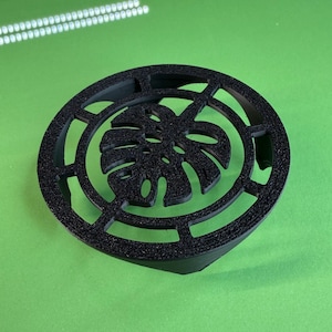 May include: A black, circular coaster with a textured surface. The coaster features a cut-out design of a monstera leaf in the center, surrounded by a geometric pattern. The coaster is set against a green background.