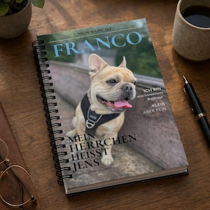 May include: A notebook with a spiral binding, featuring the name "FRANCO" in blue. The cover shows a smiling French Bulldog wearing a harness, with German text. A pen and a coffee cup are on the wooden surface.
