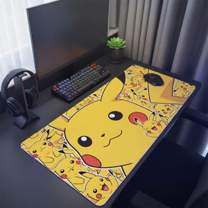 Puede incluir: Una alfombrilla de escritorio rectangular con un patrón repetitivo del personaje amarillo Pikachu con mejillas rojas, orejas con puntas negras y varias expresiones faciales. La alfombrilla está sobre una superficie oscura con un monitor, teclado y ratón.