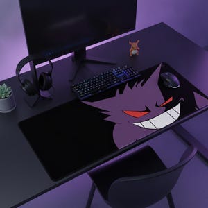 Pode incluir: Um tapete de secretária retangular com um grande gráfico roxo do Gengar com olhos vermelhos e um sorriso branco largo num fundo preto. O tapete está colocado numa secretária escura com monitor, teclado preto, rato e auscultadores.
