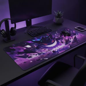 May include: A rectangular desk mat featuring a vibrant purple and black graphic illustration of a stylized creature with a wide grin, surrounded by sharp geometric shards. The mat is positioned on a dark desk surface with a computer monitor and mouse.