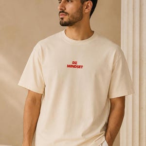 Peut inclure: Un t-shirt à manches courtes de couleur crème avec le texte "DZ MINDSET" imprimé en rouge au centre de la poitrine. Le vêtement présente une encolure ronde et une coupe décontractée, fabriqué dans un tissu opaque et uni à la texture lisse.