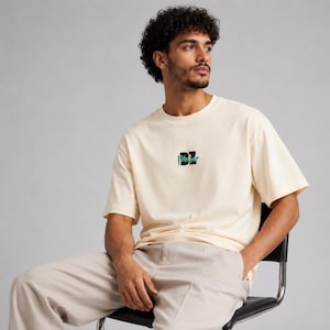 Peut inclure: Un t-shirt à manches courtes de couleur crème avec une encolure ronde et un motif central sur la poitrine. Le motif se compose du texte noir "DZ" avec un contour vert et une petite ligne d'accent verte en dessous, sur un fond uni.