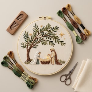 Puede incluir: Un bastidor de bordado con una escena de la natividad, un árbol y estrellas, rodeado de hilos de bordar verdes, marrones y dorados, un porta agujas, tijeras y tela.