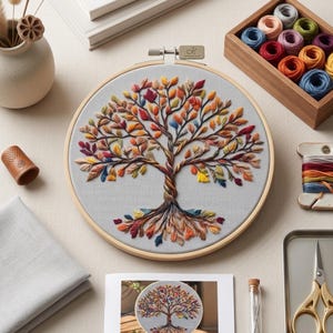 May include: A circular wooden embroidery hoop containing light gray fabric with a hand-stitched tree design. The tree features a twisted trunk and branches adorned with multicolored leaves in shades of red, orange, yellow, blue, and purple.