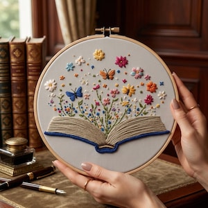 Puede incluir: Un bastidor de bordado circular de madera que contiene un panel de tela gris claro. El bordado representa un libro abierto con páginas beige y lomo azul, del cual emergen flores coloridas y dos mariposas en tonos rosa, amarillo y azul.