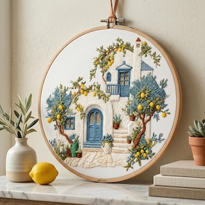 Pode incluir: Arte bordada em bastidor com uma casa de estuque branco com portas e janelas azuis, rodeada por limoeiros. A obra de arte inclui a palavra "ARTISAN". O bastidor é suspenso por uma tira de couro.