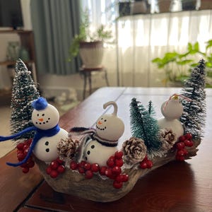 May include: A decorative centerpiece featuring three white snowman figures on a piece of driftwood. The snowmen wear scarves and are surrounded by miniature green bottle brush trees, small brown pinecones, and clusters of bright red berries.