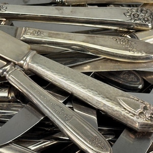 May include: A collection of antique silver butter knives and spreaders. The handles feature various decorative patterns, including floral and geometric designs. The blades are smooth and reflect the light.