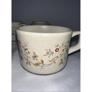 Op de afbeelding: Een gespikkelde beige keramische mok met een delicaat bloemmotief met bruine, gele en groene ranken en bloesems. De mok heeft een brede, ronde vorm, een stevig handvat aan de rechterkant en een glad, lichtgekleurd binnenoppervlak.