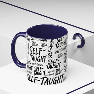 May include: A white ceramic mug with a dark blue interior and handle. The exterior surface features a repeating pattern of the words "SELF-TAUGHT" printed in a black, brush-stroke style font oriented in various directions across the entire mug.