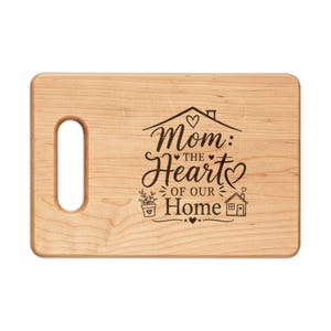 Puede incluir: Tabla de cortar de madera clara con un asa en el lado izquierdo. La tabla presenta el texto grabado "Mom: The Heart of Our Home" en una escritura decorativa, junto con ilustraciones de corazones y casas.