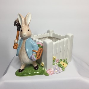 May include: A ceramic planter featuring Peter Rabbit holding a shovel, wearing a blue jacket, and standing next to a white picket fence. The planter has a basket with the words "Peter Rabbit" and is decorated with flowers.