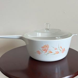 May include: A white ceramic saucepan with a long handle and a clear glass lid. The exterior of the pot features a floral pattern with orange and pink petals and green leaves. The lid has a clear glass knob handle centered on the top surface.