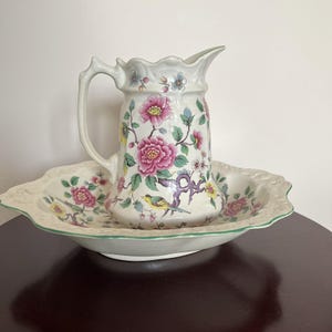 May include: A cream-colored ceramic pitcher and matching wash basin set featuring a colorful floral pattern with pink peonies, green leaves, and a small yellow bird. The pitcher has a scalloped rim and a curved handle, while the basin has a wide, embossed edge.