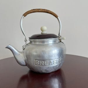May include: A brushed aluminum grease container shaped like a teapot with a spout and a handle wrapped in brown wicker. The lid is dark brown with a white knob, and the word GREASE is embossed in a rectangular frame on the front of the body.