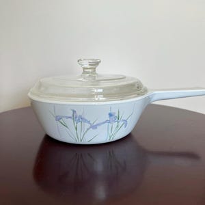 May include: A white ceramic skillet with a long handle and a clear glass lid. The exterior of the pan features a delicate floral pattern with light blue and purple iris flowers and green stems painted against a clean white background.