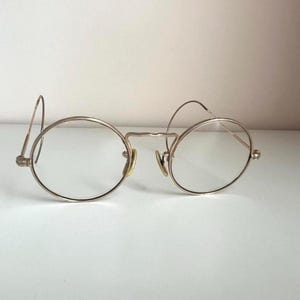 May include: A pair of round, wire-rimmed eyeglasses with clear lenses. The frames are a light gold color, featuring a bridge and nose pads. The temples curve around to rest on the ears. The glasses are set against a plain white background.