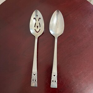 May include: Two silver-toned metal spoons with a polished finish. One spoon features a slotted bowl with a decorative cutout pattern, while the other has a solid bowl. Both handles have vertical ridges and a floral embossed design at the base.