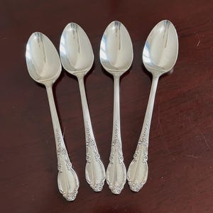 May include: Four silver-toned metal spoons arranged in a row. Each spoon features a polished oval bowl and a slender handle decorated with embossed floral and scroll patterns at the base. The metal surface has a reflective, metallic finish.
