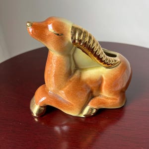 May include: A ceramic figurine of a reclining gazelle or antelope with a glossy orange-brown glaze. The animal features metallic gold accents on its nose, eyes, hooves, and curved horns, with an opening on its back for use as a small planter or vase.