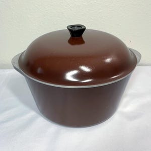 May include: A deep, round, dark brown cooking pot with a matching domed lid. The pot features a smooth, glossy finish and a small, black, circular knob centered on the lid. The rim of the pot and the lid edge are finished in a metallic silver tone.