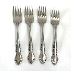 May include: Four silver forks with ornate handles. The forks have tines and a decorative pattern on the handles. The forks are arranged in a row against a white background. The silverware is likely for a formal dining setting.