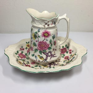 May include: A vintage cream-colored pitcher and basin set. The set features a floral design with pink peonies, yellow flowers, and green leaves. The basin has a scalloped edge with a green trim. The pitcher has a handle and a decorative rim.