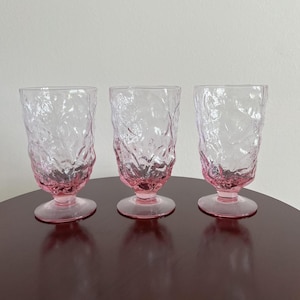May include: Three matching pink glass goblets featuring a textured, crinkled surface pattern. Each glass has a rounded bowl, a short stem, and a circular base. The pink color is more saturated at the bottom and fades toward the top rim.