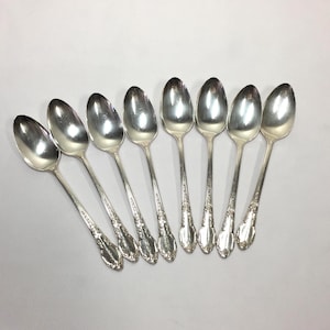 May include: A collection of eight silver spoons arranged on a white surface. The spoons have a classic design with ornate detailing on the handles and a polished, reflective finish. The spoons are likely for dining.