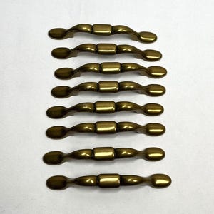 May include: A set of eight identical brass-toned metal drawer pulls arranged in a vertical column. Each pull features a smooth, curved design with a central rectangular block and rounded ends, showcasing a polished metallic finish throughout.