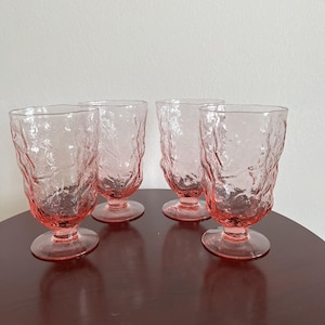 May include: A set of four pink glass tumblers with a textured, uneven surface and a footed base. Each glass features a translucent, light pink hue and a distinct, organic, crinkled pattern across the exterior, standing on a circular pedestal.