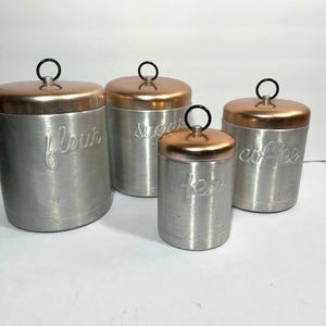 May include: A set of four cylindrical metal canisters with brushed silver bodies and copper-toned lids. Each lid features a black circular handle. The canisters are embossed with the cursive words flour, sugar, tea, and coffee on their sides.