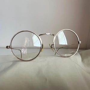 May include: A pair of round eyeglasses with thin gold-toned metal frames and clear lenses. The glasses feature a curved bridge, adjustable nose pads, and slender temples that extend outward. The design is simple and symmetrical with circular rims.