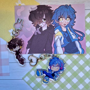 Könnte beinhalten: Ein Illustrationsdruck und zwei Acryl-Schlüsselanhänger mit Anime-Charakteren. Der Druck zeigt zwei Charaktere, einen mit braunen Haaren und einen mit blauen Haaren. Die Schlüsselanhänger zeigen die Charaktere in Chibi-Form.