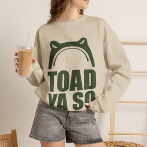 Könnte beinhalten: Ein beigefarbenes Sweatshirt mit Rundhalsausschnitt und einer dunkelgrünen Grafik, die den Umriss eines Krötenkopfes sowie den Text "TOAD YA SO" in fetten Blockbuchstaben zeigt. Das langärmelige Kleidungsstück hat gerippte Bündchen.