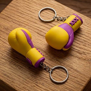 Può includere: Due portachiavi a forma di guantoni da boxe gialli e viola con anelli argentati. I guantoni presentano dettagli viola e cuciture. I portachiavi sono esposti su una superficie di legno.