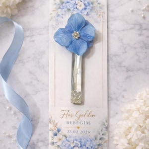 Puede incluir: Una flor azul con un centro de perlas está unida a un soporte rectangular transparente. El soporte está en una tarjeta con el texto "Hos Geldin BEBEGIM 25.02.2026" y un diseño floral. También hay una cinta azul claro y perlas.