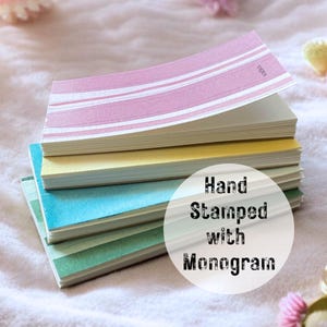 May include: A stack of colorful stationery with a pink, yellow, blue, and green color scheme. The top sheet is pink with white stripes. The text "Hand Stamped with Monogram" is in a white circle.