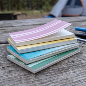 May include: A pile of notebooks with covers in various colours and patterns. The top notebook has a pink striped cover, followed by yellow, blue, and green. The notebooks are on a weathered wooden surface, with pens visible in the background.