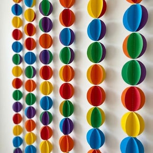May include: A vertical garland consisting of multiple strands of colourful paper circles. Each strand features a repeating pattern of circular paper discs in vibrant shades of red, orange, yellow, green, blue, and purple, all stitched together in a line.